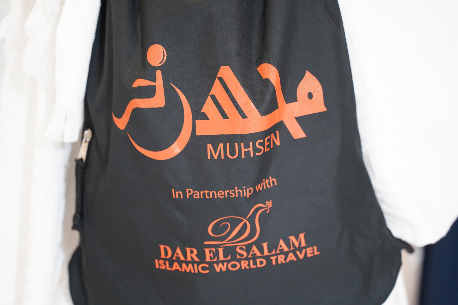 Black drawstring backpack with orange lettering saying "MUHSEN in partnership with Dar-el Salam Islamic World Travel."