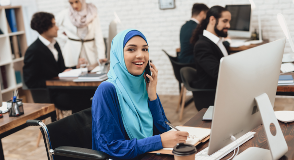 girl in a blue hijab talking on the phone while sitting at a computer.